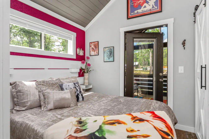 Cozy modern bedroom with plush gray bedding and colorful face-print throw, magenta accent stripe under a narrow horizontal window, vaulted ceiling, glass patio door opening to a wooded yard and small deck, framed art and vase of flowers.