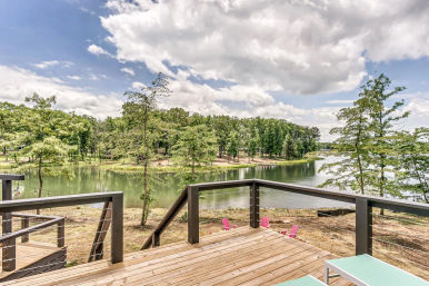 Lakefront wooden deck with cable railing overlooking a calm tree-lined lake and pink Adirondack chairs on the shore under a partly cloudy sky