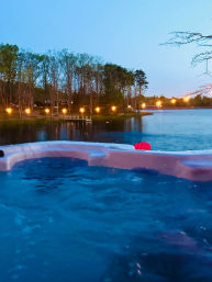 Cozy lakeside hot tub at dusk with bubbling blue water in the foreground, twinkling string lights along a tree-lined shore and a small dock reflecting on the calm lake.