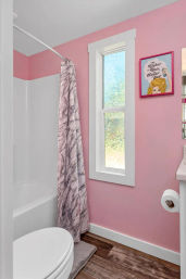 Small pink bathroom interior with white-trim window, tub-shower with marble-pattern curtain on a curved rod, white toilet, wood-look flooring and playful framed art on the wall.