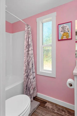 Small pink bathroom interior with white-trim window, tub-shower with marble-pattern curtain on a curved rod, white toilet, wood-look flooring and playful framed art on the wall.
