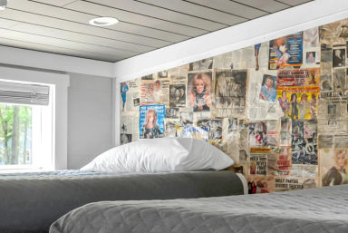 Cozy loft-style bedroom with two twin beds, gray quilted bedding and white pillows, slanted ceiling and small window with leafy view, playful vintage magazine and newspaper collage accent wall