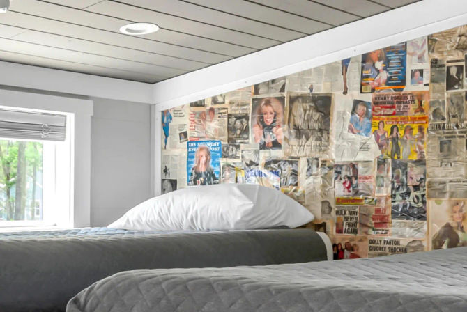 Cozy loft-style bedroom with two twin beds, gray quilted bedding and white pillows, slanted ceiling and small window with leafy view, playful vintage magazine and newspaper collage accent wall
