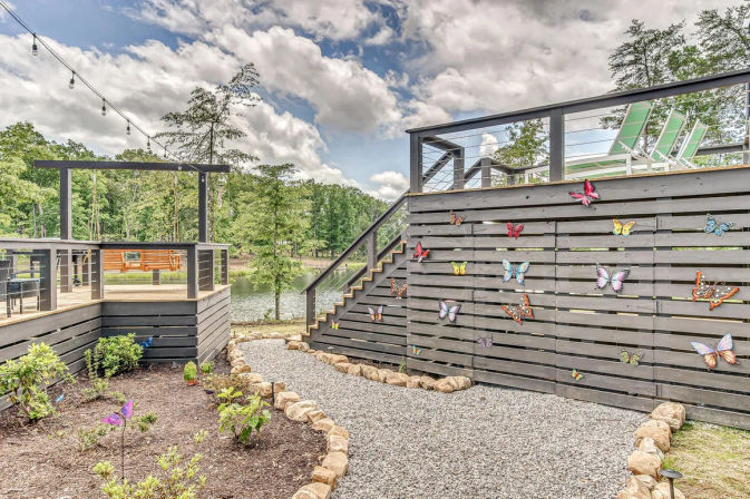 Lakefront gray wooden deck and pergola with string lights, gravel garden path edged with stones, and a playful butterfly‑adorned privacy wall overlooking a wooded pond.