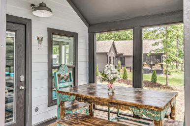 Screened porch with a rustic reclaimed-wood dining table, distressed turquoise chairs and bench, glass vase of white daisies, and a green backyard with cottage-style houses