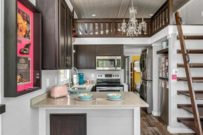 Compact tiny-house kitchen with marble island and colorful bowls, dark wood cabinets, stainless appliances, pink retro toaster, stacked washer-dryer, loft with crystal chandelier and open wooden stairs