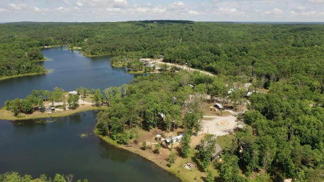 Aerial view of a forested lakeshore campground with winding blue lake, cabins and RV sites tucked among green trees, dirt roads and a central firepit — peaceful rural nature retreat