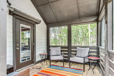 Cozy screened wooden porch with two gray patio armchairs, small round side tables, a colorful striped rug and a glass door, overlooking a wooded backyard view.