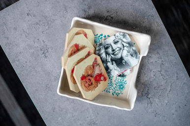 Hexagonal vintage portrait coasters stacked in a white ceramic dish with a blue floral pattern on a gray stone tabletop — one black-and-white smiling glamour portrait and several colorful pin-up style images.