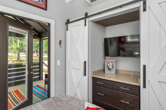 Cozy bedroom with white sliding barn doors opening to a niche with a wall-mounted TV and dark wood dresser topped by a decorative retro-portrait book; glass door leads to a screened porch with a colorful striped rug and wooded yard views.
