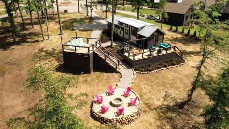 Aerial view of a tiny house on a wooded lot with an elevated wooden deck and turquoise seating, connected by a stone path to a circular stone fire pit surrounded by bright pink Adirondack chairs.