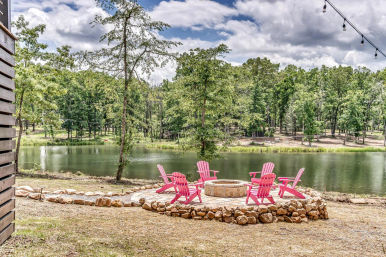 Lakeside fire pit area with bright pink Adirondack chairs arranged around a stone fire pit on a rock-edged patio, overlooking a calm tree-lined pond under a partly cloudy sky with string lights overhead.
