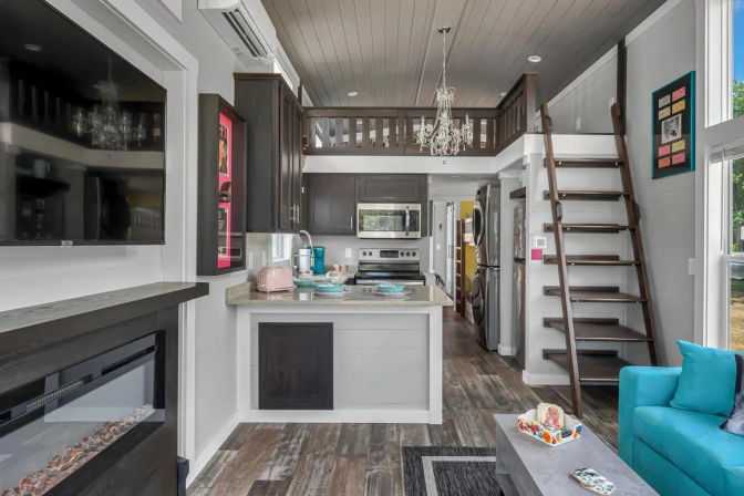 Cozy modern tiny house interior, open-concept kitchen and living area with dark wood cabinets, stainless appliances, center island, crystal chandelier, loft accessed by wooden ladder, turquoise sofa, wall-mounted TV above electric fireplace, and distressed wood floors