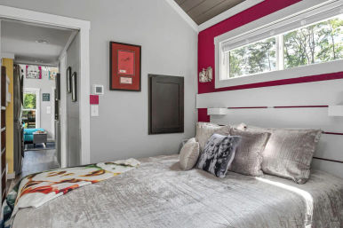 Cozy modern compact bedroom with gray walls and magenta accent, silver bedding and decorative pillows, horizontal clerestory window and view into an adjoining living area.