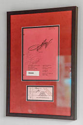 Framed memorabilia: red script cover page with bold black signatures and a matching ticket stub mounted on a red mat inside a slim black frame on a light gray wall.