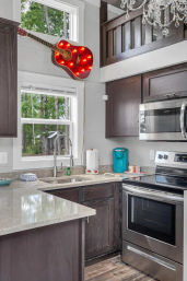 Modern kitchen with dark wood cabinets, quartz countertops and stainless steel range, teal coffee maker, and a red light-up guitar wall decor above a window overlooking a wooded backyard.