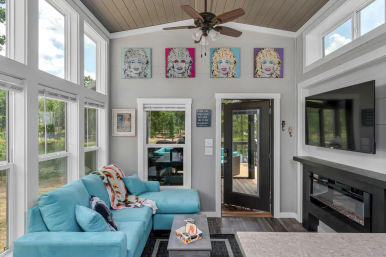Bright lake-view sunroom with floor-to-ceiling windows, turquoise L-shaped sectional, colorful pop-art portraits on a gray wall, wall-mounted TV above a modern electric fireplace, ceiling fan and glass door leading to a deck.