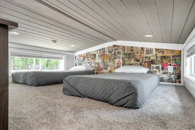 Cozy low-ceiling attic loft bedroom with two gray floor mattresses on plush carpet, quilted bedding, a playful vintage magazine-collage slanted wall, and long windows with a forest view.