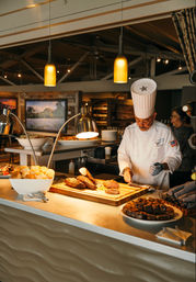Chef in white uniform carving roast at a restaurant buffet carving station under warm pendant lights, with bowls of dinner rolls and dessert squares on the counter.