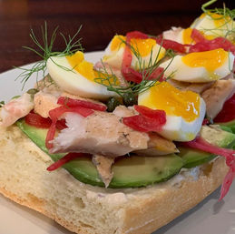 Gourmet open-faced avocado toast on crusty bread topped with sliced avocado, flaked smoked fish, soft‑boiled egg halves with runny yolk, pickled red onions, capers and dill.