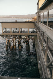 Oceanfront pier restaurant on wooden pilings with diners visible through large windows, weathered boardwalk and a seagull beside churning water