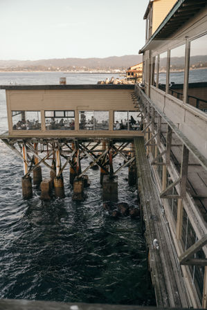 Oceanfront pier restaurant on wooden pilings with diners visible through large windows, weathered boardwalk and a seagull beside churning water