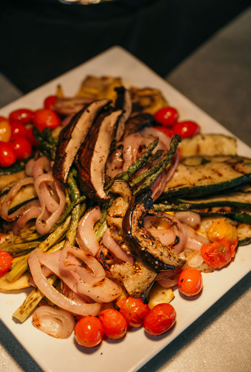 Close-up of a colorful grilled vegetable platter — charred zucchini, asparagus, portobello mushroom slices, grilled red onion and blistered cherry tomatoes on a white plate.