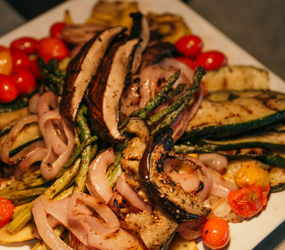 Close-up of a colorful grilled vegetable platter — charred zucchini, asparagus, portobello mushroom slices, grilled red onion and blistered cherry tomatoes on a white plate.