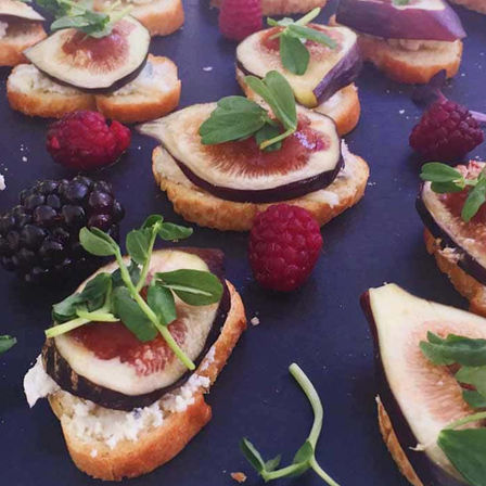 Fig crostini appetizers on toasted baguette slices with goat cheese, fresh fig slices, microgreens, and scattered raspberries and blackberries on a dark serving platter