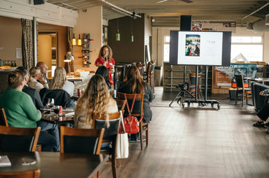 Presenter in red addressing a seated audience during a daytime workshop in a cozy restaurant-style event space with tables, drinks, and a large screen slide.