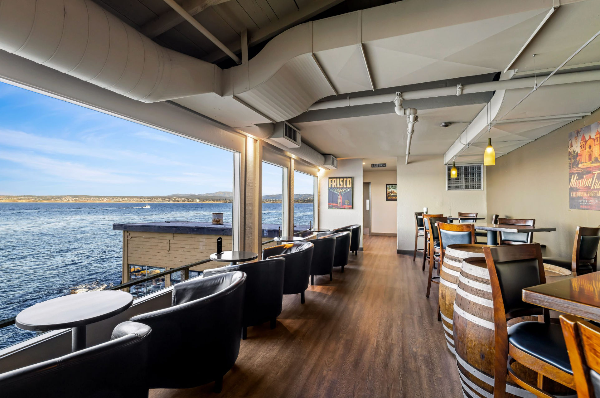 Sunlit waterfront restaurant interior with panoramic ocean view, row of black leather armchairs at window tables, wooden floors and barrel-style tables.