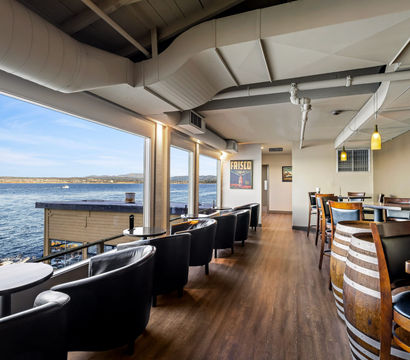 Sunlit waterfront restaurant interior with panoramic ocean view, row of black leather armchairs at window tables, wooden floors and barrel-style tables.