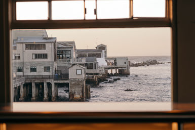 Window-framed ocean view of weathered seaside pier buildings on concrete pilings above a rocky shoreline at golden hour