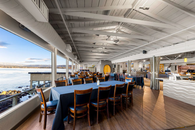 Sunlit oceanfront dining room with panoramic bay views through large windows, long blue-clothed tables, wooden chairs and white exposed-beam ceiling.