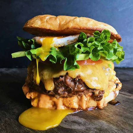 Close-up of a mouthwatering gourmet cheeseburger with melted cheddar over a juicy beef patty, crisp green lettuce and tomato, runny fried egg yolk dripping onto a toasted bun and creamy sauce on a wooden board