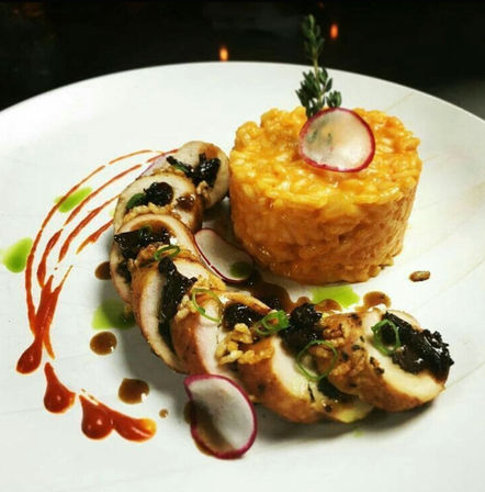 Gourmet plated entrée of sliced stuffed chicken roulade with crunchy topping, paired with a creamy yellow risotto tower, garnished with radish slices, herb sprig and artistic red and green sauce swirls on a white plate