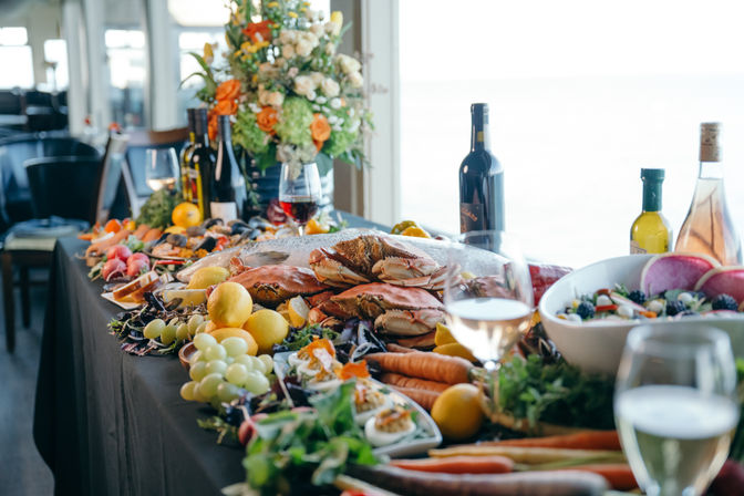 Seaside feast of whole steamed crabs piled with lemons, grapes, salads, carrots and wine bottles on a table with a floral centerpiece and ocean view.