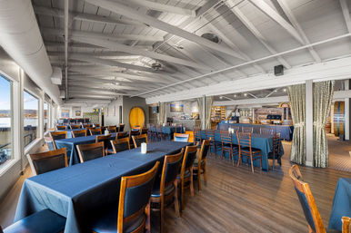 Bright waterfront event space with navy‑cloth banquet tables and wooden chairs, white exposed‑beam ceiling, large windows overlooking a harbor and rustic barrel decor