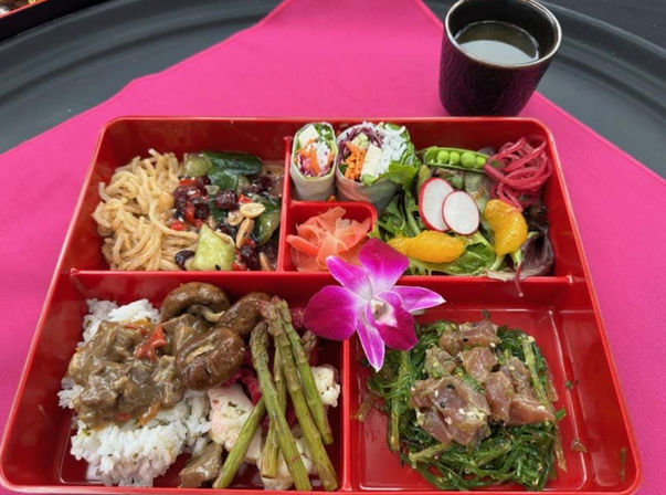 Red bento box lunch with Asian-fusion dishes: white rice topped with mushrooms and savory sauce, grilled asparagus, tuna poke on seaweed salad, cold sesame noodles, fresh vegetable spring rolls, mixed greens with radish and mandarin slices, pickled ginger, purple orchid garnish and soy dipping cup on a pink placemat.