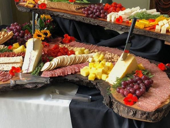 Rustic charcuterie spread on live-edge wood slabs at a buffet: cheese wedges with knives, sliced salami, round crackers, red grapes, cubed yellow cheese, cherry tomatoes, nuts, fresh herbs and edible flowers.