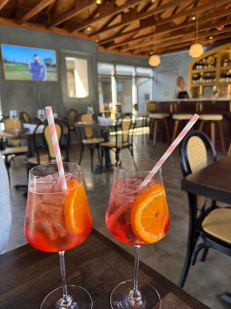 Two orange-slice spritz cocktails with ice and pink-striped paper straws on a wooden table in a sunlit bistro with rattan chairs, bar and wood-beamed ceiling in the background