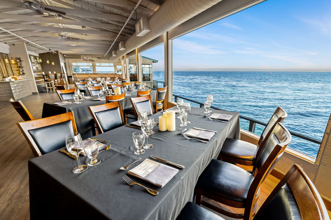 Coastal waterfront restaurant interior with panoramic ocean view, long black-tablecloth table set for dinner with wine glasses, candles and wooden chairs beside floor-to-ceiling windows