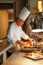 Professional chef in a tall white toque slicing roasted beef on a wooden cutting board at a warm-lit buffet carving station with heat lamps and plated sides in the foreground.