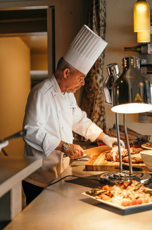 Professional chef in a tall white toque slicing roasted beef on a wooden cutting board at a warm-lit buffet carving station with heat lamps and plated sides in the foreground.