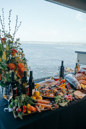 Oceanfront seafood spread with whole fish, shellfish paella, fresh vegetables and wine bottles on a table by a window overlooking a calm seaside view, bright floral centerpiece.