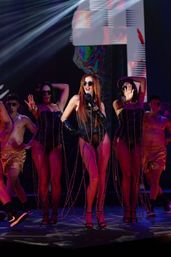 Lead singer in black bodysuit, long gloves and sunglasses performs on a neon-lit concert stage with backup dancers in fringed corsets and shirtless dancers in gold shorts, dramatic spotlights and LED backdrop.