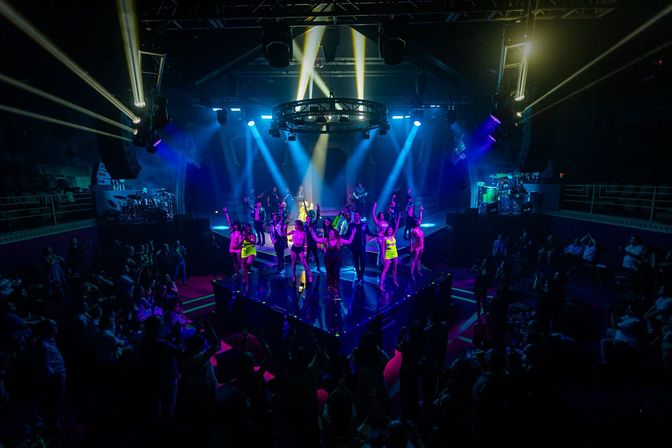 Energetic nightclub performance on a raised square stage: a troupe of dancers in bright outfits and a live band under dramatic blue and yellow spotlights, surrounded by a standing and seated audience in a dark concert hall atmosphere.