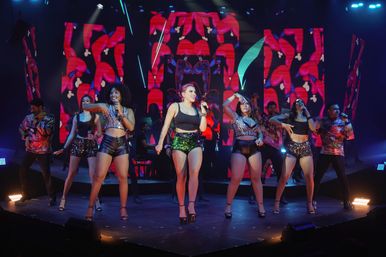 High-energy live concert with a group of singers and dancers in sparkly and leather outfits performing a choreographed routine on a lit stage with vibrant red-patterned LED screens and dynamic stage lighting.