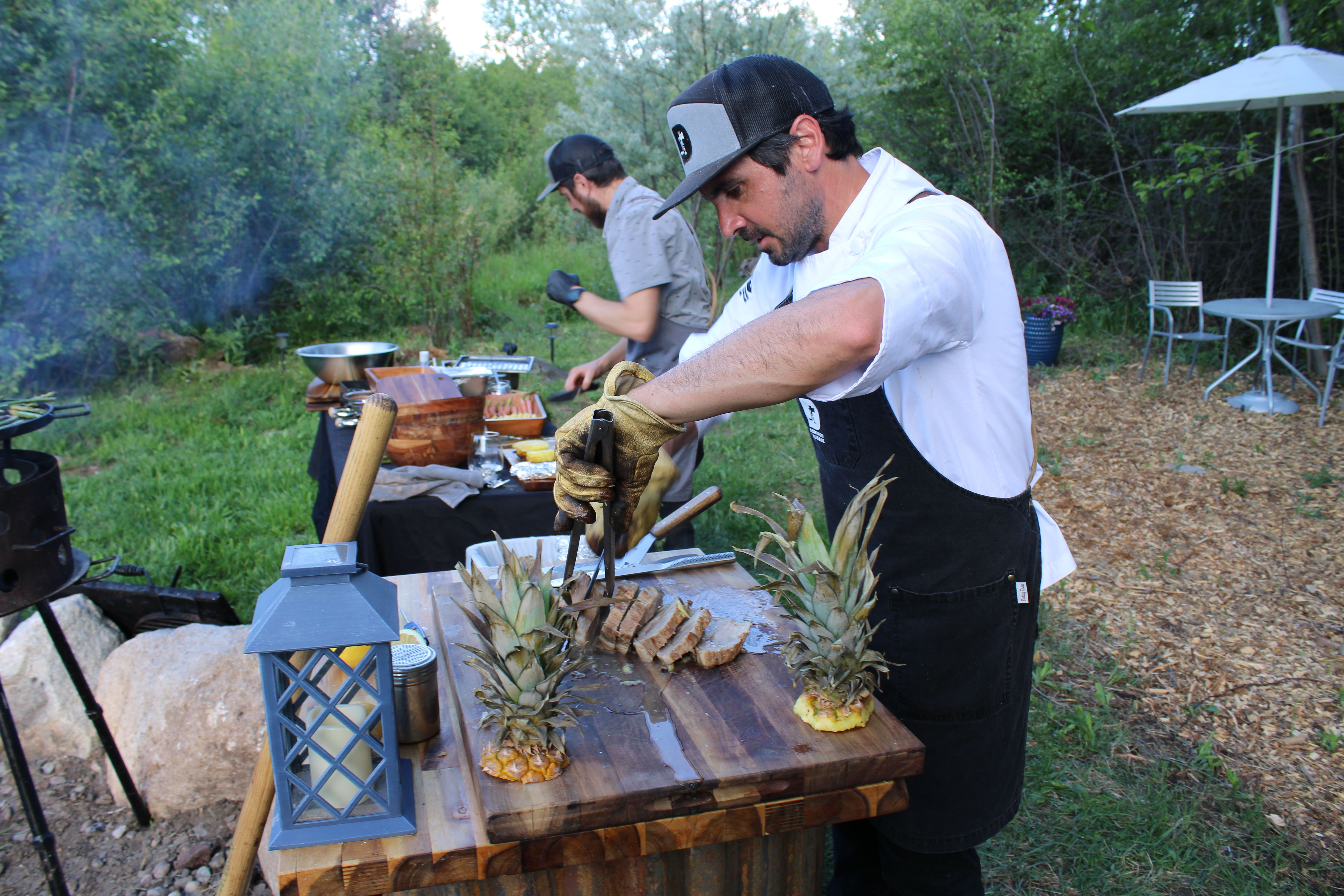 Chef in apron slicing grilled meat on a wooden cutting board with pineapple garnishes at a rustic outdoor garden barbecue prep station.