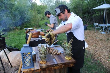 Chef in apron slicing grilled meat on a wooden cutting board with pineapple garnishes at a rustic outdoor garden barbecue prep station.